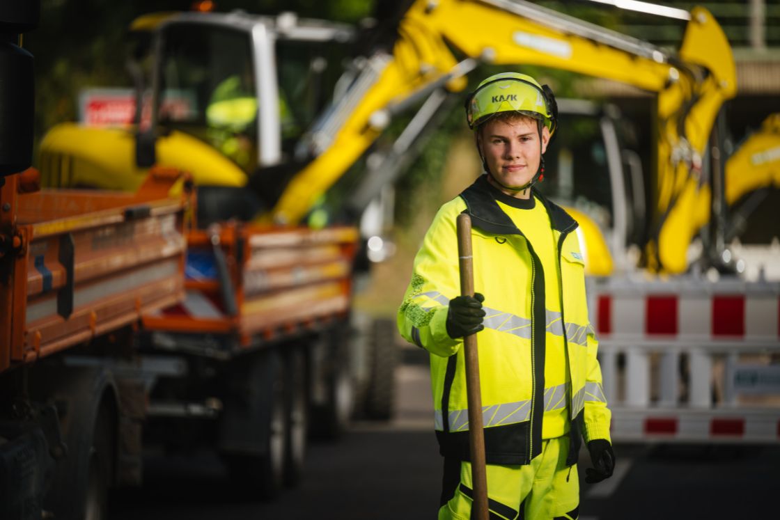 junger Mann in Arbeitskleidung mit Schaufel in der Hand auf einer Baustelle. Im Hintergrund Baustellenfahrzeuge